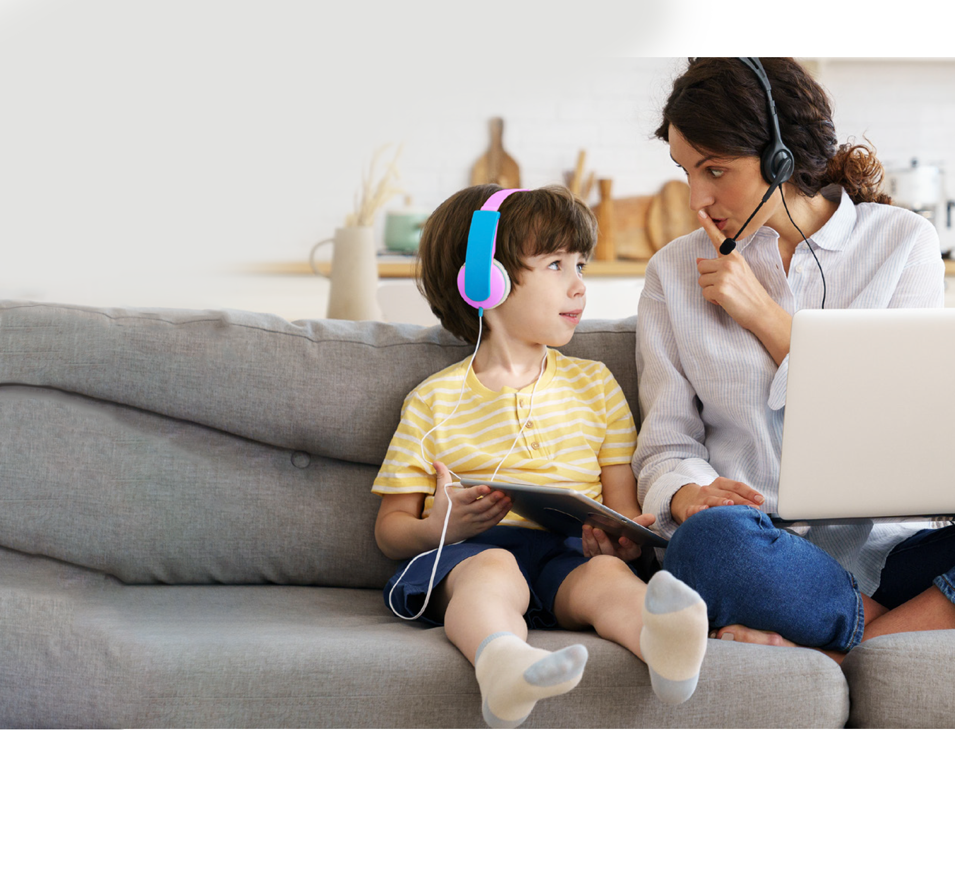 A woman on her laptop and headset is shushing her child who also has headphones on.