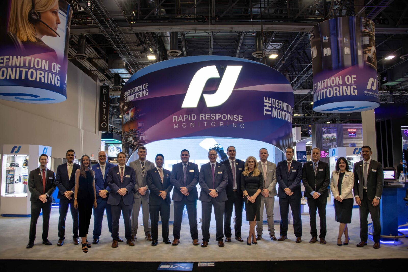 Meet Our Sales Team 8 A Group Of Professionals Standing Together In Front Of A Trade Show Booth For Rapid Response Monitoring. The Booth Features A Large Curved Display With The Company'S Logo And The Tagline &Quot;The Definition Of Monitoring.&Quot; The Team Is Dressed In Business Attire, And The Exhibition Hall Is Visible In The Background With Other Booths And Signage.